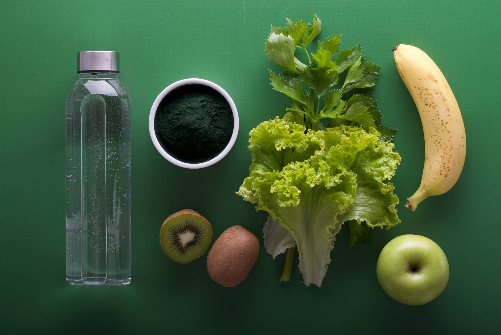 Fresh greens and fruit with a bottle of water and a bowl of spirulina powder on a green background.