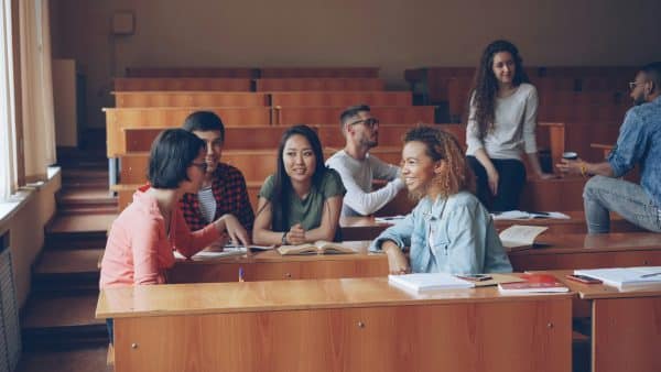 Group of students in a classroom chatting and studying around wooden desks with notebooks and papers