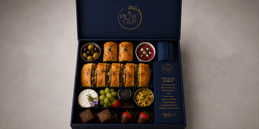 09- The Picnic Club Gourmet Picnic Basket | OTS News - Southport Deluxe navy gift box opened to reveal an assortment of wraps, olives, hummus, cheeses, fruit, chocolates, and desserts labeled The Twilight Basket by Picnic Club.