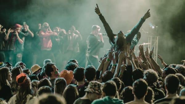 Crowd at a concert with a person being lifted above the audience and reaching upward amid stage lights and smoke.