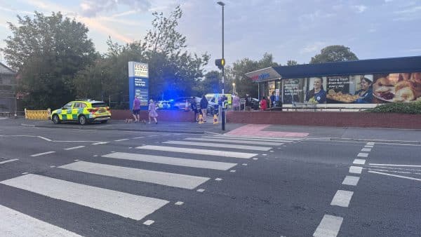 Tesco Express storefront with pedestrians and a police car with flashing blue lights in the parking lot behind a crosswalk.