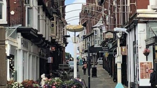 Narrow pedestrian street in a busy shopping district, brick buildings on both sides, colorful flower displays, and a few people walking in the distance.