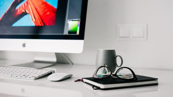 Desk with an iMac, wireless keyboard and mouse, notebook, glasses, and a mug on a clean white surface.