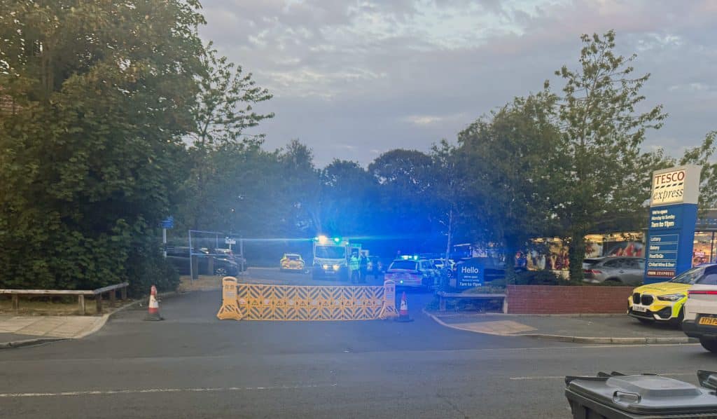tesco2 | OTS News - Southport Tesco Express storefront behind police barricades with flashing blue emergency lights at dusk.