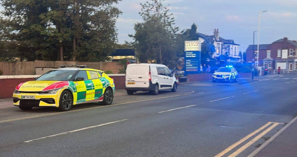 tesco3 | OTS News - Southport Emergency scene on a suburban street with a police car flashing blue lights, a white van, and a colorful emergency vehicle in the foreground near Tesco Express signage.