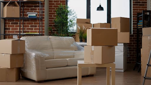 Living room filled with cardboard boxes and a wrapped beige sofa, brick walls in the background.