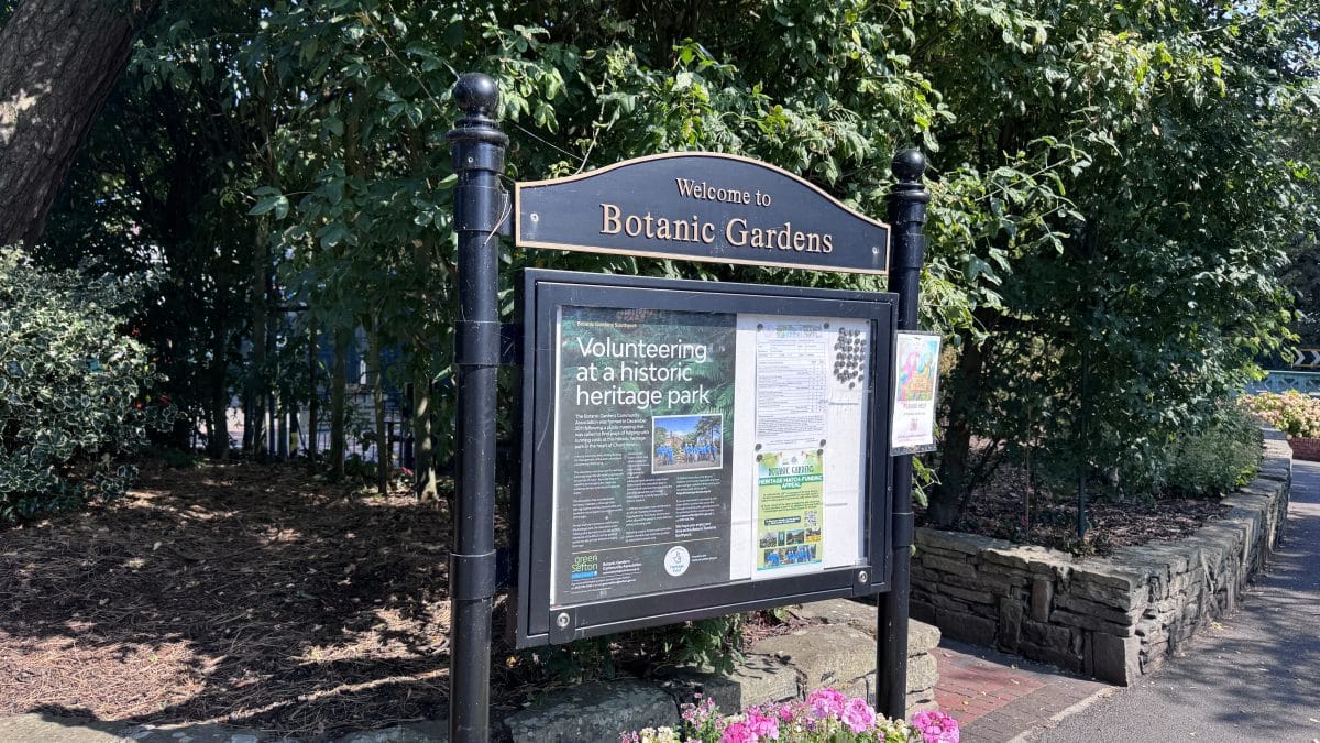 Sign reading 'Welcome to Botanic Gardens' with an information display, black posts, and lush greenery nearby.