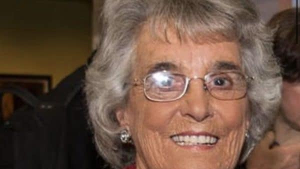 Smiling elderly woman with short gray hair and glasses, wearing a red outfit and pearl necklace at a social event.