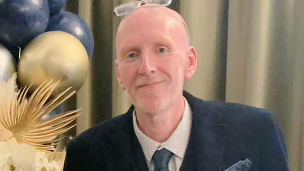 Smiling man in a navy blazer with a light shirt and tie, wearing glasses on his head, balloons in the background.